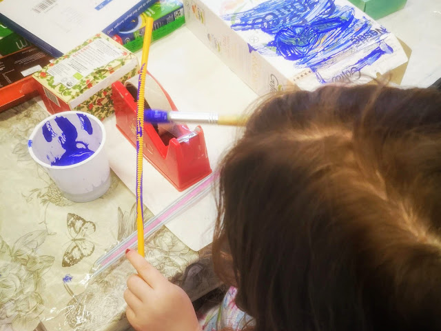 A close up image of a child painting a plastic straw with blue paint. The paint is in a plastic cup. On the table is a patterned table cloth, a red tape dispenser and a collection of cardboard boxes and product packaging to be used for junk modelling.