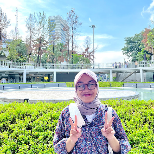 Foto seorang wanita (alias aku) di area terbuka Taman Literasi Martha Tiahahu. Sebuah gedung pencakar langit terlihat di latar belakang