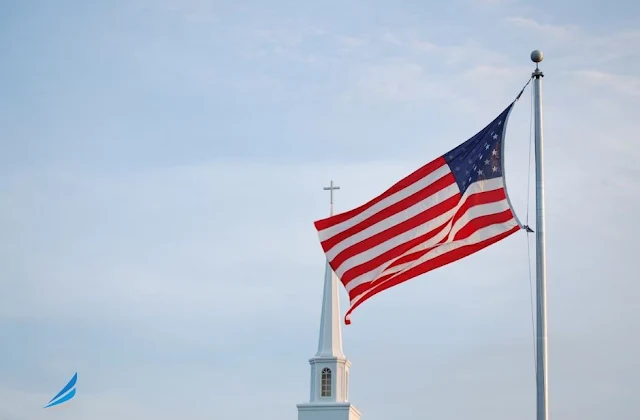 American flag waving on flagpole with white church steeple and cross visible in background against blue sky via unsplash