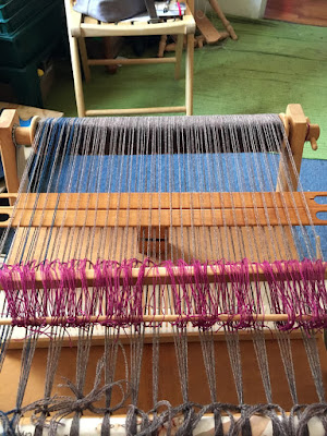A small wooden rigid heddle loom warped with grey and teal, with two flat wooden stick shuttles holding some of the warp threads up. A thin dowel holds the ends of dozens of magenta cord loops, and is laid on the near side of the heddle, while the loops disappear behind the heddle.