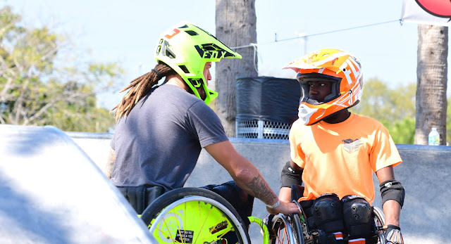 A main in a grey shirt, riding in a bright green manual wheelchair and wearing a matching motorcycle helmet, is shown talking to a young man who is also a manual wheelchair user. He rests his hand on the wheel of the other person's wheelchair. The young man is wearing a bright orange shirt. His wheelchair and motorcycle helmet match his shirt.