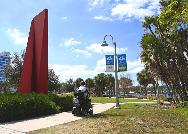 Kyle drives past a red, modernist steel art piece, titled "America." It is located between two sidewalks that veer off in different direction. It's surrounded by a patch of green, waist-height ferns. To his right is a patch of grass and a light pole, which has to, blue banners that have the logo for Riverwalk.