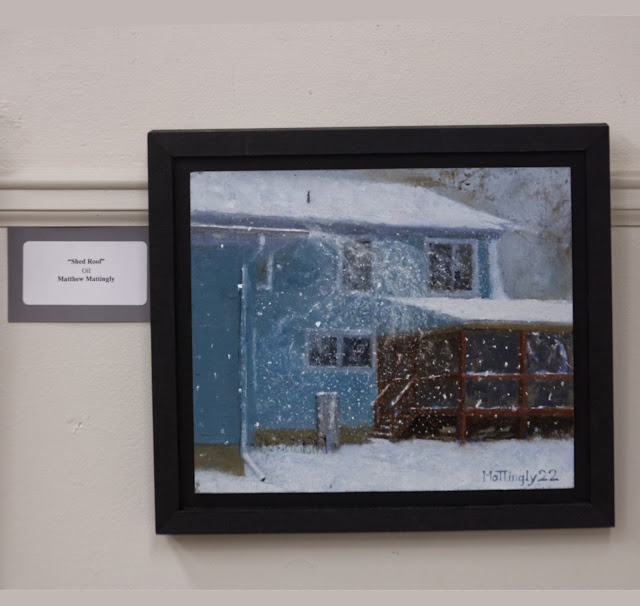 Oil painting of house with snow blowing off of roof, hanging on gallery wall. Label on left side says "Shed Roof" Oil Matthew Mattingly