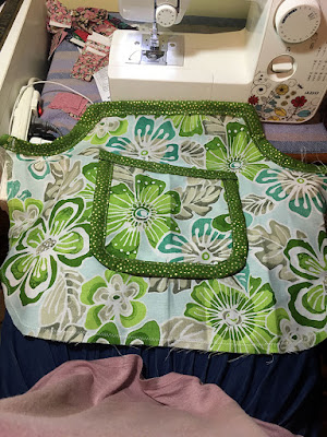 A stacked set of pockets, made of green, turquoise, and grey tropical floral patterned fabric and bound in light lime-green fabric with densely scattered gold dots. The smaller pocket is square with rounded bottom edges, bound all the way around, and centered on the larger, rectangular piece with quarter-circle cutouts from the top corners, and rounded bottom corners. The larger piece is bound only across the top edge, with a dense white zigzag stitch following the other three edges about half an inch in.