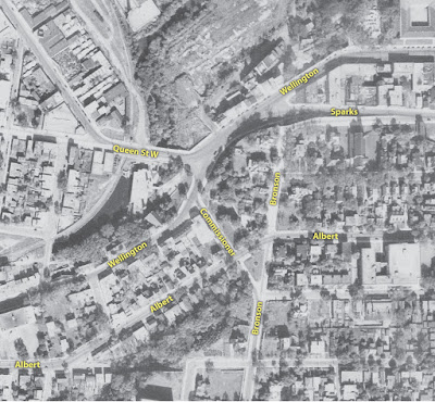 Crop of a 1944 black and white aerial photograph showing the area around Wellington and Commissioner, with yellow street labels I've added on Wellington, Sparks, Queen St W, Commissioner, Bronson, and Albert. Wellington crosses Queen St W and Sparks Street in a stretched X intersection (almost a double Y), and off Wellington at the south end of this forks Commissioner, which crosses Albert connects in to Bronson at Slater. Unlike today, traffic coming north on Bronson can connect into downtown on Wellington Street via Commissioner.