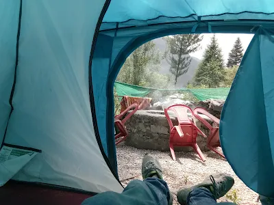 View from inside a camping tent showing a relaxed person’s legs stretched out, with an outdoor firepit surrounded by plastic chairs, trees, and a misty mountain backdrop.