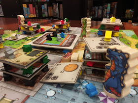 A similar view of the board, this time from the point of view of the blue monster with the green and red monsters visible in the background. However, this time, some of the cardboard 'floors' of the buildings have been knocked off the stack onto the board, and the meeples have likewise been knocked around.