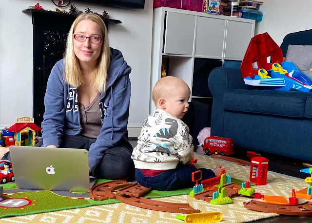 A mum on a MacBook sitting on the floor trying to work and surrounded by toys and a baby