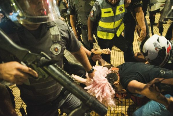 Um policial armado segura uma mulher pelos cabelos. Ela está agachada e ao redor, nota-se outros policiais.