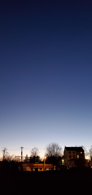 A photo of distant trees, a house, and streetlights, with the darkening evening sky above.