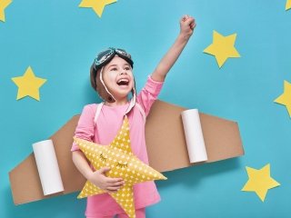 A girl wearing cardboard rocket wings and holding a star, pretends to soar into space.