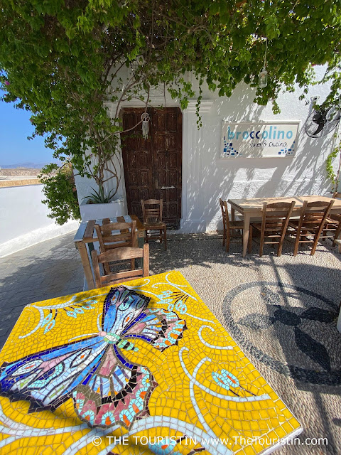 A bistro table tiled with a bright yellow butterfly tabletop in front of a brown wooden entrance door of a whitewashed house on a pebble stone floor under a wine-covered gazebo.