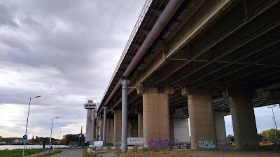 The underside of a huge motorway bridge. There are additional steel columns and a narrower bridge attached to the motorway bridge here.