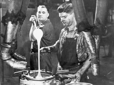 Black and white image of two men working in a glass factory. One holds a rod with molten glass, which he is pouring into a mould, while the other man holds a paid of shears, about to snip off the molten glass.