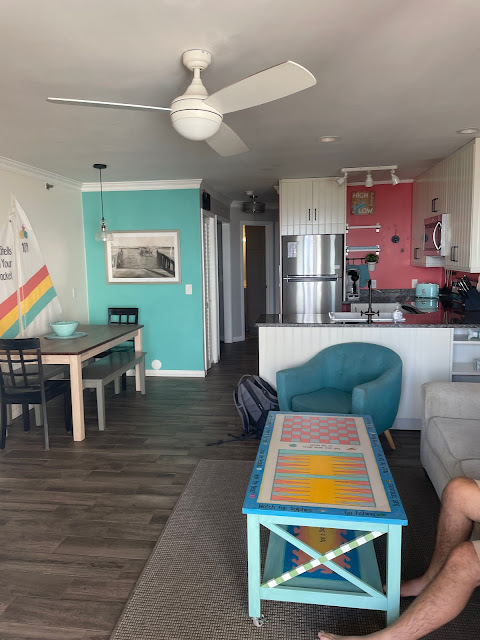 This is a view of the airbnb from the front door. In front, on the right hand side of the room is the couch with the end table. to the back, right of the room is a table with two chairs and two benches. There are two walls in view of the dining table - one is aqua and one is white with decor on the walls. On the back right of the photo is the kitchen with a burnt orange wall. The appliances are stainless steel. There is a hallway in the middle back of the room which leads to the bathroom, pocket bedroom and the master.