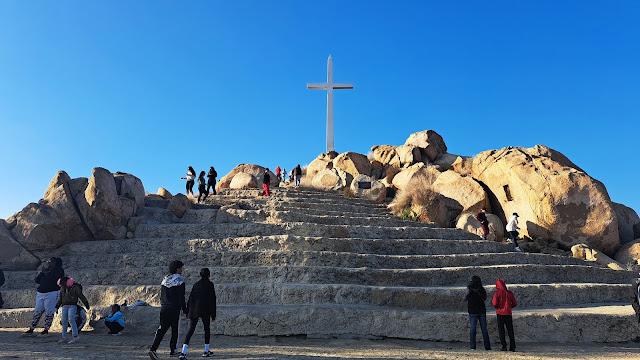 Cross atop a hill nestled amidst boulders with massive stairs leading to it. People stare and some walk to it and some are near it.