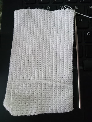 A rectangle of white cotton thread crocheted into a rectangle lies against the black background of a laptop keyboard. The steel crochet hook lies next to the white rectangle. The rows seem to form vertical stripes (since I've rotated the project 90 degrees in this picture, sorry!)