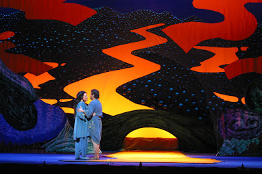 Photo of an opera: A man and a woman dressed in blue tunics and matching blue pants embrace in front of a colorful backdrop depicting a golden river running down a hill towards the front of the stage, with a bridge crossing it. The backdrop is almost cartoonlike.