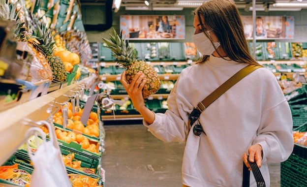 Image of woman wearing a mask, holding a pineapple, and doing grocery shopping.
