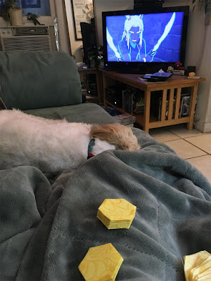 A shot of a person's lap under a fuzzy grey-blue blanket, with stacks of yellow English paper-pieced hexagons on their knees and a small white dog at their feet, and a still from The Dragon Prince on the tv.