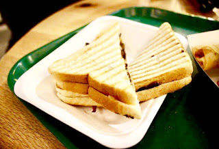 Two pieces of a white triangular sandwich with black grill marks scorched on it with a dark brown meat interior and white oozing interior to represent the cheese on a white square plate on a light background. 