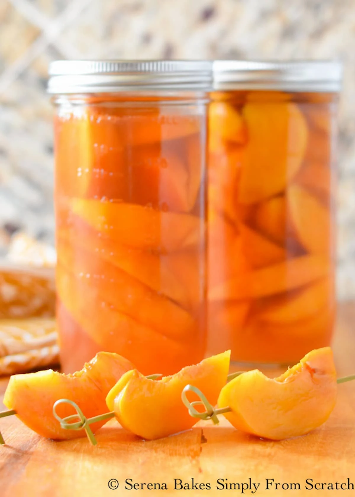 Two jars of Whisky Peaches with three skewered peaches in front to garnish a drink.