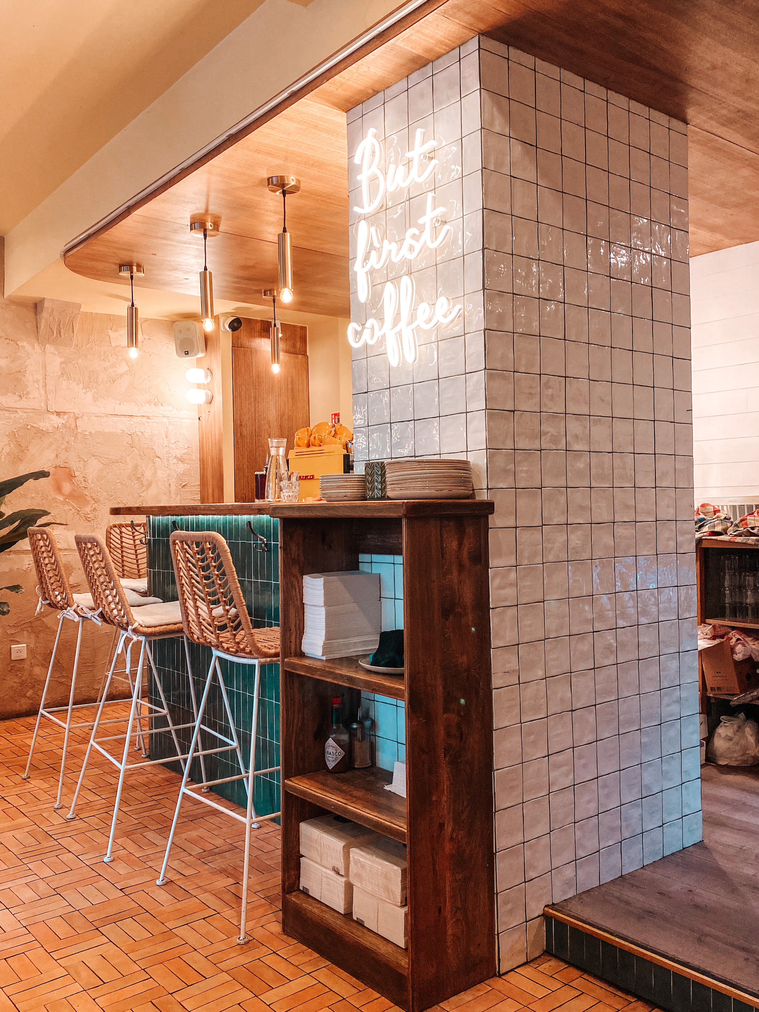 intérieur du restaurant immersion à paris avec des chaises hautes en rotin et un mur de brique blanc sur lequel est écrit but first coffee avec des néons allumés