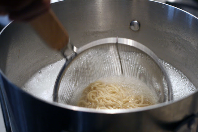 A wire mesh strainer containing the noodles in a pot of hot water.