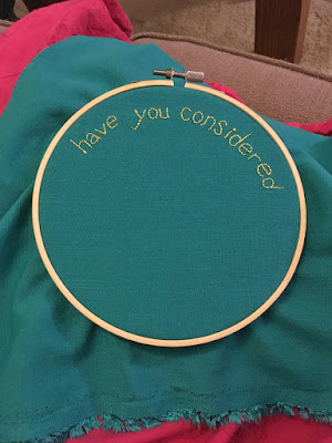 A plain wooden embroidery hoop with a dull silver screw, holding turquoise fabric embroidered with "Have you considered" in small yellow backstitch around the upper edge of the circle.