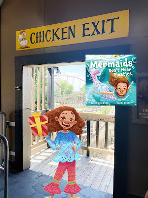 A cartoon girl with red hair and light brown skin, wearing a blue shirt and red striped pants and holding a wrapped present, stands in front of a real life door with a rollercoaster in the background. Above the door is a yellow sign that reads "Chicken Exit"