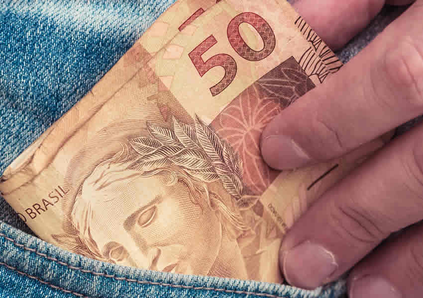 Imagem mostra uma mão colocando algumas notas de 50 reais em seu bolso da calça.