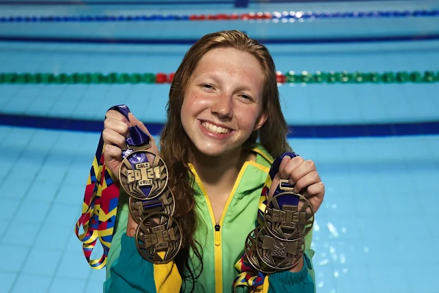 Stephanie posa para foto sorrindo com suas medalhas. Ela segura as medalhas com suas duas mãos (quatro em uma mão e três na outra) e veste um agasalho verde do Time Brasil