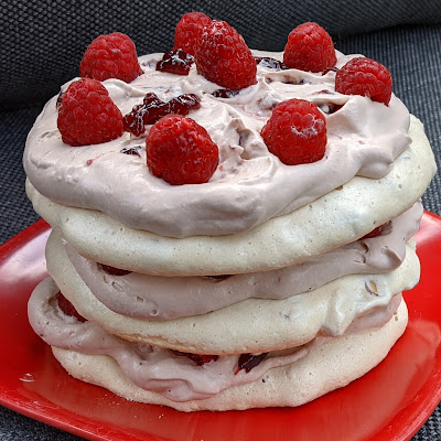 three discs of creamy white dacquoise on a red plate, alternating with layers of rosy brown ovaltine whipped cream, raspberry jam, and fresh raspberries