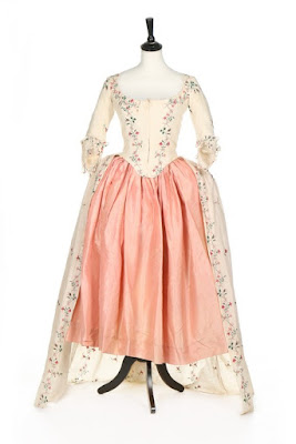 A sale image of a white, delicately floral-sprigged 18th-century gown with simple cuffs and open skirt, styled with a shell-pink petticoat on a white display mannequin.