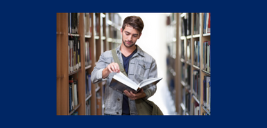 A young man with brown hair and a beard and wearing a navy blue T-shirt and denam jacket is holding a book and he is surrounded by libary shelves.