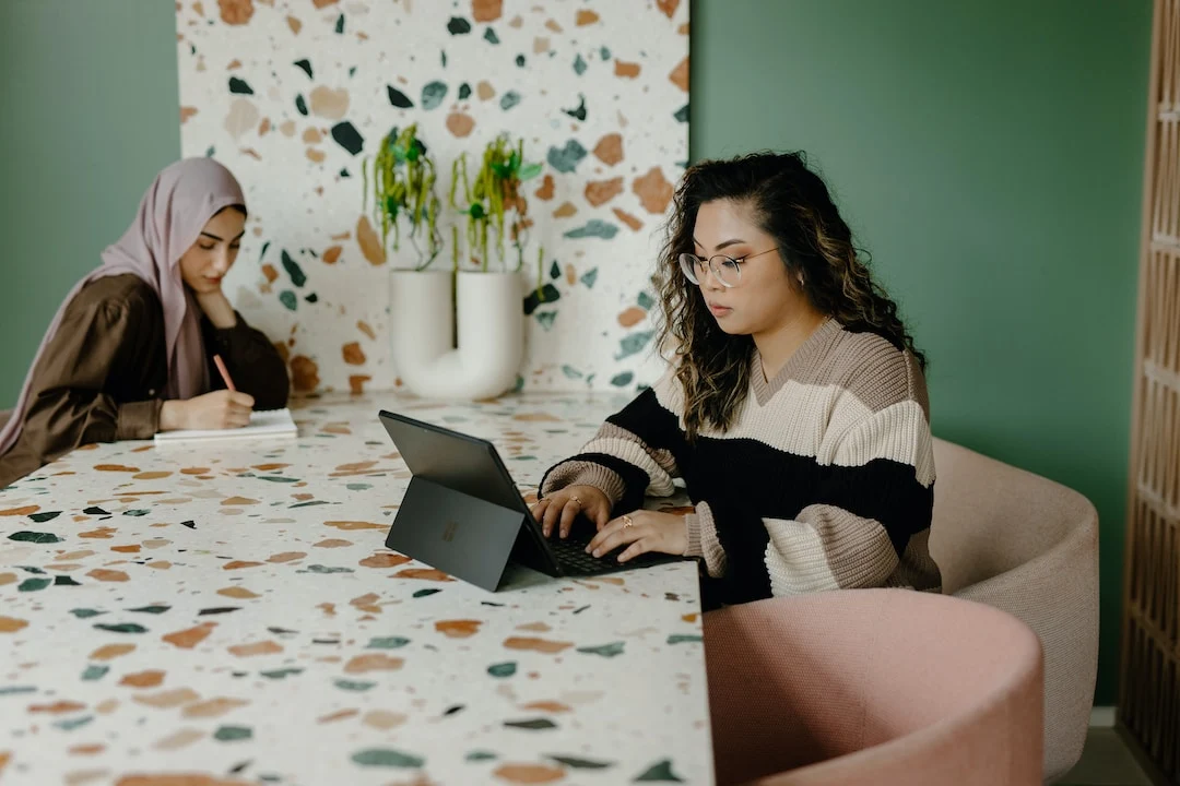Two girls sitting in front of each other one wearing hijab and other working on a tablet wearing t-shirt and glasses photo