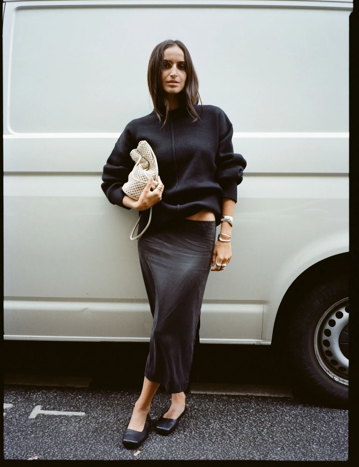 Woman with medium dark brown hair standing in the street in front of a white van wearing an oversized black fleece jacket, a black satin mini skirt, black ballet flats, chunky statement silver bangle bracelets and carrying a beige crochet macrame bag