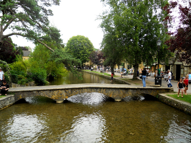 Bourton on the Water, A lovely little town in Cotswolds. Bourton on the Water is the a tidy Cotswolds town that sits on both the banks of the river Windrush. It is a major tourist stop and is often referred to as the Venice of the Cotswolds because of the river running through it and its low, stone bridges. This was the prettiest of all the Cotswolds towns that we visited on this trip.Our tour bus dropped us at the parking lot and we had to walk to the centre of the town where the river flows. On the way we came across several trademark Cotswolds cottages and this close called Foxes Close. Close in British Sense means a narrow alley or pathaway.People relaxing on the banks of the river Windrush. Your would've seen similar scenes on handouts distributed by various builders of apartment complexes. They lead the buyers to believe that the lifestyle in the apartment complexes will be as laid back and chilled out as in some of these British Towns.A low stone bridge on the river Windrush. You find numerous cafes and Souvenir shops on both the banks of the rivers. Children playing freely in the shallow river and fishermen idling around with their fishing rods doing their work for them are part of the common weekend scene in the town. An alley with yellow cottages on both the sides. Tidy grey streets and yellow walls are in stark contrast with each other. People picnicking on the banks of the river. You'll see people of all age-groups having fun. The best part of this lifestyle is that no one judges the others too much. Old people are entitled to having as much fun as the youngsters. My Friend Hannah. She was constantly attracted to shops selling antiques and vintage stuff. Me and Kelsey were equally interested/disinterested in everything so we had no issues in following her around. Cotswolds was the best part of this trip and Bourton on the Water was the best town we visited in the Cotswolds.Cotswolds, Bourton on the Water, River Windrush, UK, Travelling Camera, Travel