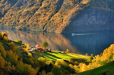 A house by a green field on a fjord with colorful forests all around it. There is a boat moving on the water.