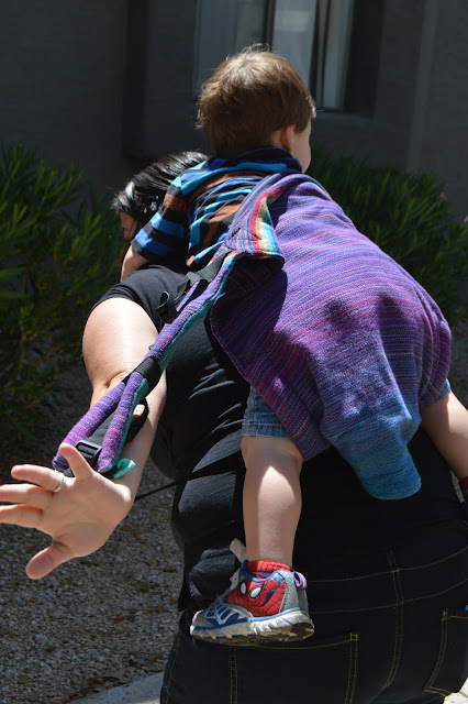 Image from behind as woman slides her arm into the shoulder strap with one arm as she holds baby's arm with her other hand