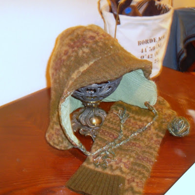Earflap cap with brim, constructed from repurposed sweaters. Outer layer is of boiled wool, patterned in shades of olive and brown, with the brim a solid olive ribbing. Inner layer is of light green ribbed material. With the hat is a pair of arm-warmers, cut from sleeves of the sweater that forms hat's outer layer. In background: a canvas tote holds thread and pieces of hats for sewing.