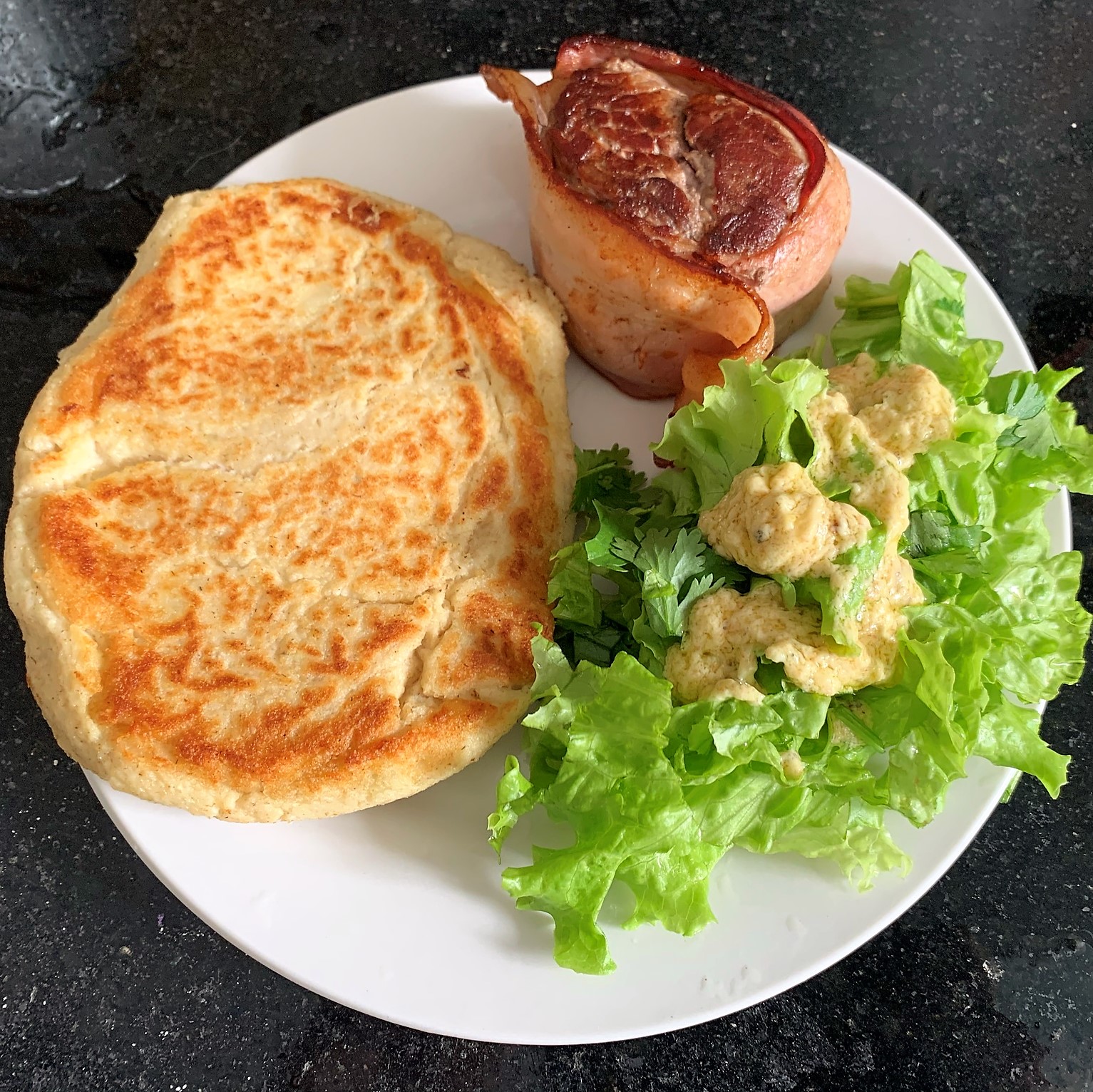 Foto de Bolinho de Batata, medalhão de filet de mignon com bacon e salada com molho de maionese num prato branco em cima de mármore preto