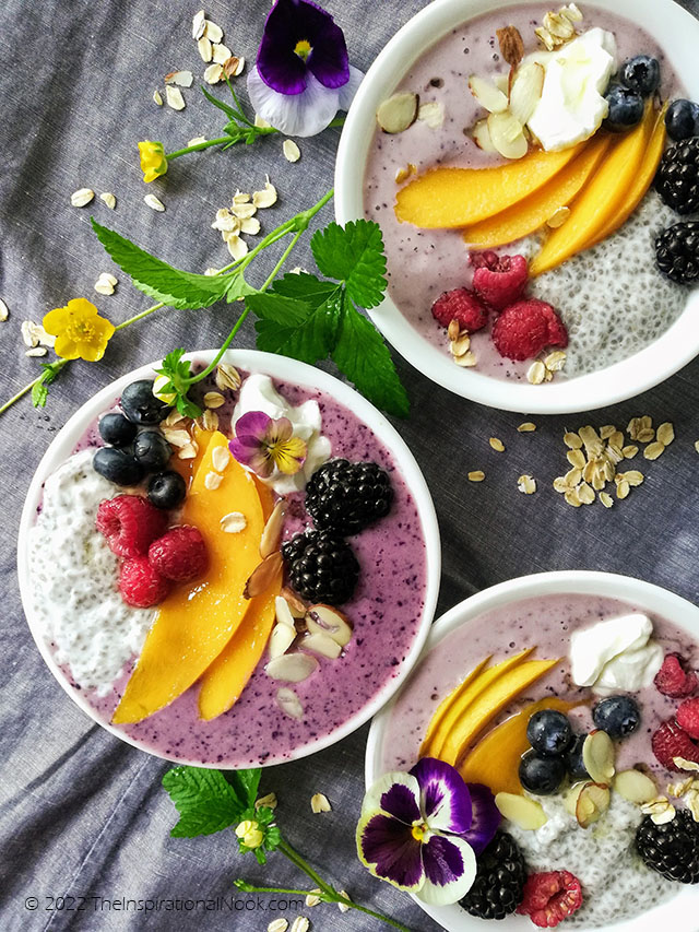 Fruit bowl with sliced mangoes, raspberries, blueberries, chia, purple smoothie, purple and yellow pansy flowers