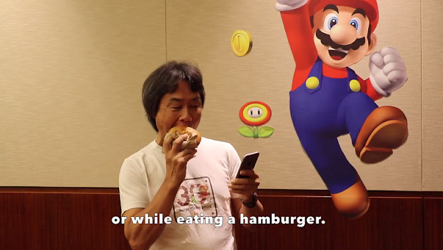 Shigeru Miyamoto eating hamburger with one hand playing Super Mario Run