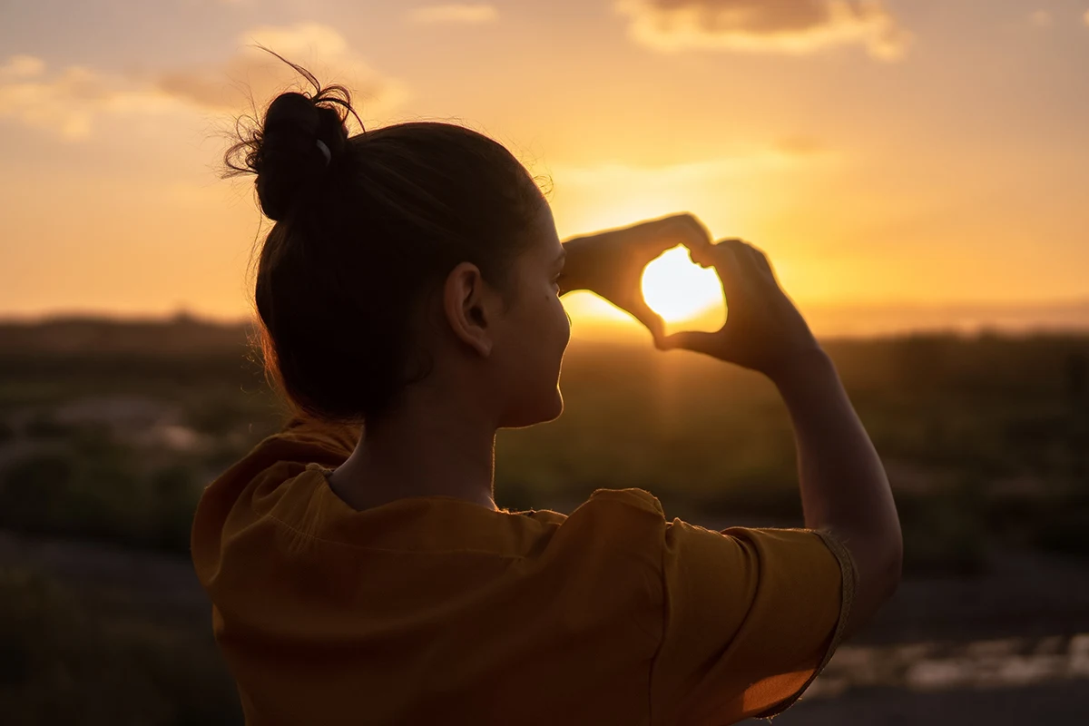 woman turned towards the sundown showing heart with her fingers