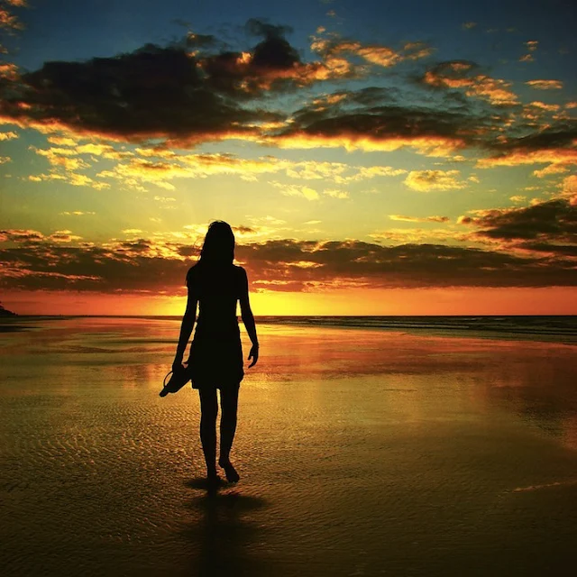 The silhouette of a person walking barefoot on a wet, reflective beach at sunset, holding shoes in one hand. The sky is ablaze with vibrant orange and yellow hues near the horizon, fading into dark blues and purples overhead, with dramatic clouds reflecting the golden light.