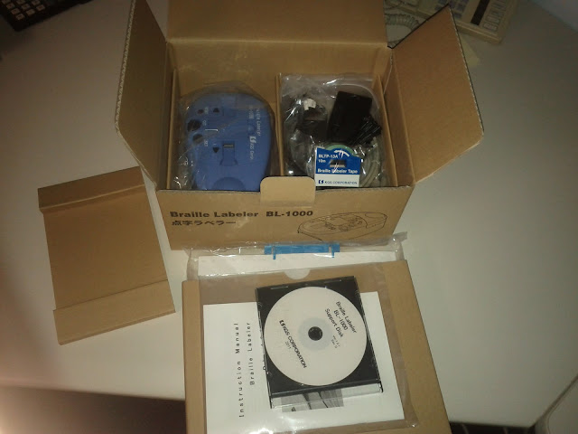 Now upon the same off-white desk, the small brown box is open and inside is a blue oval object which is the Braille Labeler, some black wires and white Braille tape, extra brown cardboard, a cd to install the program for the labeler with a white label on it, an instruction manual with a white cover and another instruction manual sticking out of another brown box that was inside the first box and contains the manual in Braille and has a blue plastic binding.