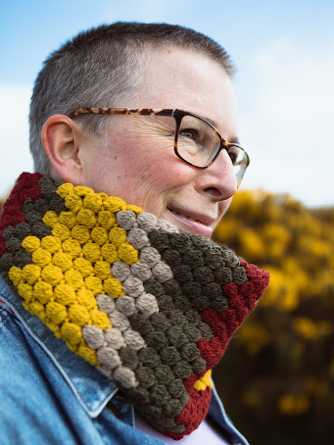 Head and shoulders shot of a woman wearing a crocheted cowl in autumnal colours of ochre, burgundy, beige and green