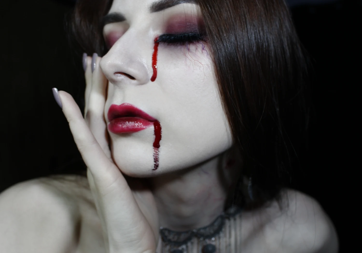 a close-up of pale woman's face with a dramatic vampire makeup look and false blood tears for Halloween.