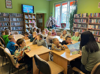 Wnętrze biblioteki. Pani bibliotekarka siedzi naprzeciwko grupy przedszkolaków i czyta im książkę Dobry Dinozaur. W tle widzimy regały z książkami, regał z czasopismami na którym stoi włączony telewizor. Na telewizorze wyświetlony jest obrazek z dinozaurami rysunkowymi i napisem 26 lutego Dzień Dinozaura.